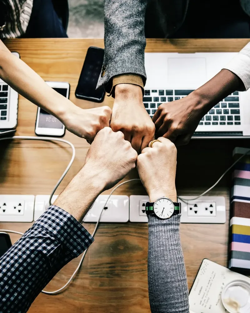 Varias manos chocan los puños sobre una mesa de trabajo con portátiles y cuadernos, vista desde arriba.